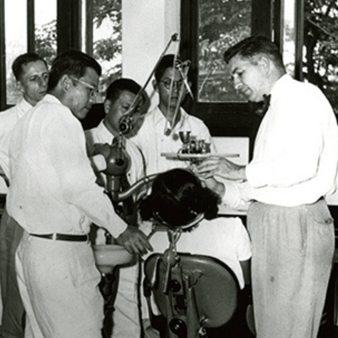 A black and white photo of Dr. Volker (white man in white button up and slacsks) showing others in professional attire sental tools over a dental chair in a 1960s dental clinic setting
